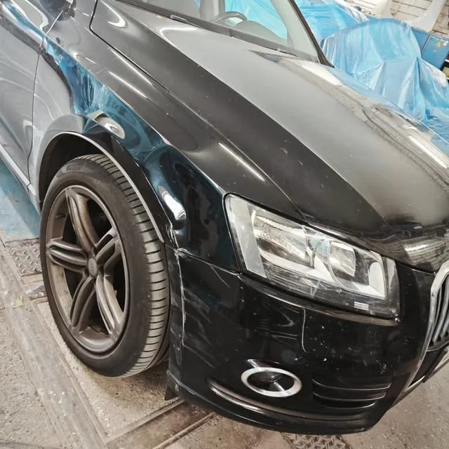Schwarzer Audi mit beschädigtem vorderem Kotflügel in der Werkstatt, dahinter blaue Abdeckfolie